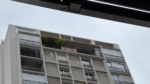 High flat building. A gray interface with a balcony full of plants. From the handrail hanging a sign that does not read any concerts in Spanish. It is written in large red letters against a white background.