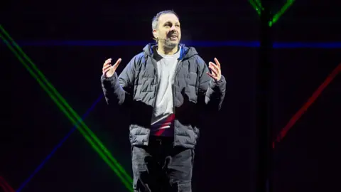 Steven Frayn stands on stage with his hands gesturing he is a man with short brown-grey hair. He is wearing black cargo trousers, a grey jumper and a black puffer jacket. Behind him are coloured lights.