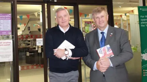 Essex County Council Russell Quirk, wearing a dark-coloured fleece, and Peter Harris in a grey suit with a blue tie, smile directly at the camera. They are holding books and standing outside the glass sliding doors of a library.