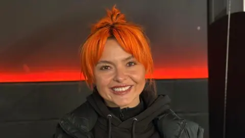 A woman, Rosie Webber, with bright orange hair smiling at the camera, wearing a black hoodie. She is inside a dimly lit gym.