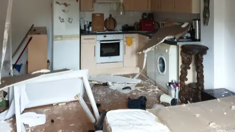 A small kitchen with white appliances and pale wood cupboards. The ceiling has collapsed and left debris across the room. A white table is on its side with broken legs.