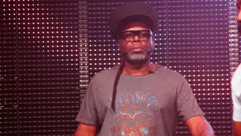 Jazzie B is standing in front of a wall of lights as he performs. He is wearing thick square framed glasses, a large black hat and a grey top with blue and orange sequins on. He has long dark hair that has been braided over one shoulder and grey and dark facial hair.