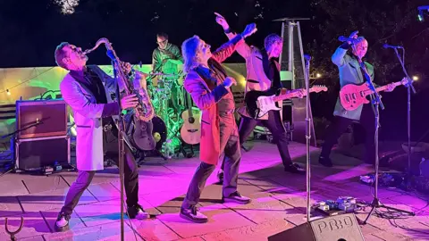 Showaddywaddy performing in the garden, pointing to the sky as they dance in teddy boy outsits, lit pink from the lights