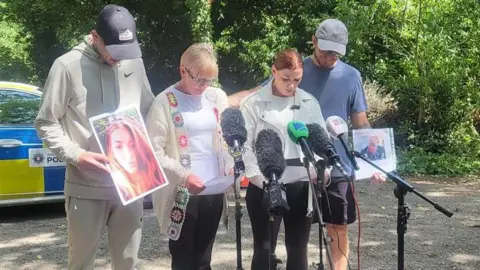 Four people standing at microphones in front of a woodland during a press release. Two women stand in the middle with a man on either side. The are holding pictures of a missing woman who has red hair. There is a police car parked behind them.