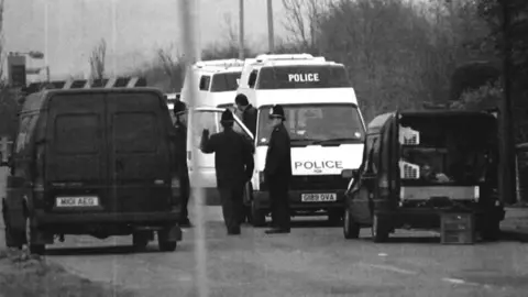 PA Media A black and white image of two police officers wearing tall hats and overcoats standing on a road near four vans. Two vans are white and two dark-coloured. The word "POLICE" is written twice on the front of one white van.