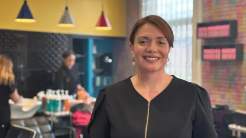 Victoria has brown hair tied back and is wearing a black top with a gold zip and gold hoop earrings. She is standing inside a hairdresser's salon smiling at the camera. In the background a woman is washing a customer's hair in a sink.