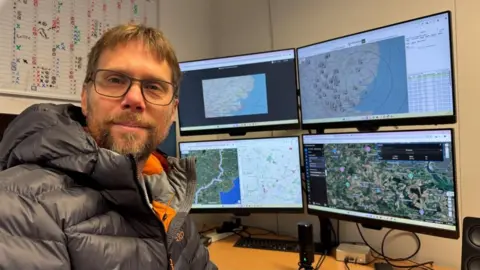 Andrew Turner/BBC Rowley Cory-Wright is wearing a puffa jacket, which is charcoal on the outside, and grey and orange on the inner lining. He is wearing glasses, has an auburn beard and moustache and medium length hair. He is sitting at a desk with four computer screens behind him, showing maps and drone images of survey areas between Norwich and Tilbury