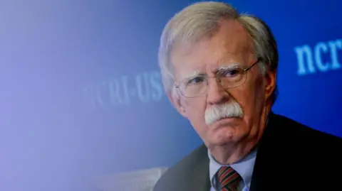 John Bolton at an event in August. He has a serious expression on his face, grey hair, a grey moustache and wire-rimmed glasses. He's wearing a black jacket, blue shirt and red and blue striped tie. There is a bright blue background.