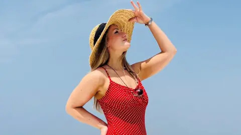 Hannah Howie, who has long, blonde hair and is wearing a red dress with white spots, strikes  a pose against a clear blue sky. Her right hand is on her hip and he left hand is tipping the top of her straw hat.