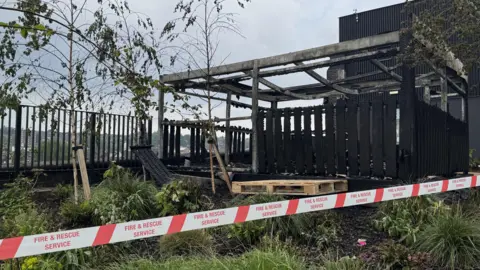 Red and white fire and rescue service tape is wrapped around the scene. In the background is a charred wooden structure that has turned completely black. A patch of shrubs and trees around the affected structure appear largely untouched.