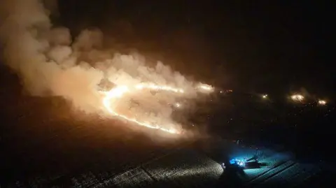 Dan Jessup An aerial night-time image showing a large grass fire burning across a field near Stone Cross. Flames form a long curved fire front with thick smoke rising into the air. Blue emergency vehicle lights are visible near the edge of the field.