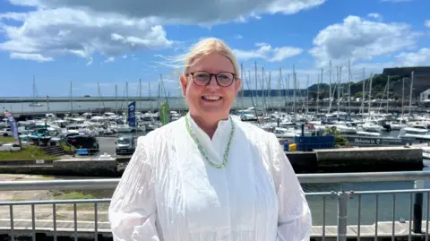 Dr Audery Cameron smiling at the camera. She is wearing a white top and green necklace. She is stood in front of Queen's Anne Battery where there are docked boats.