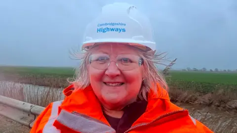 Mousumi Bakshi/BBC Lorna Dupre is stood in the middle and is wearing a white hard hat which says Cambridgeshire Highways, and is wearing an orange Hi-Vis jacket. She has short grey hair and is smiling. 