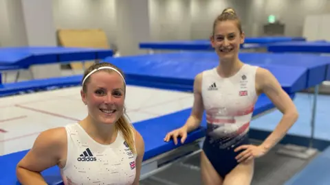 Supplied At the bottom left of the photo is Laura from the chest up, wearing a Team GB and a headband. On the right of the photo is Bryony Page in a Team GB leotard and her hair in a bun. Both are leaning  on a trampoline. 