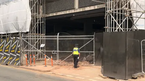 A police officer with their back to the camera close to a road is standing near silver gates and there are vertical silver beams behind the gates and to the right of them on the photo. The next storey is partly visible at the top of the image.