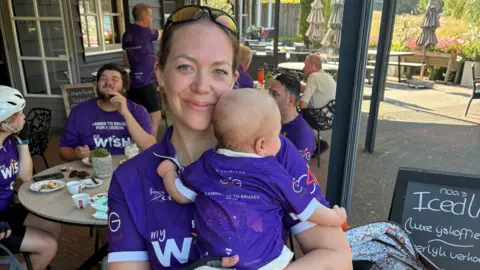 Contributed A mother holding a baby smiles at the camera. She wears a purple cycling jersey with the baby wearing a matching top. She has sunglasses resting on top of her head. There are people sat around tables in a cafe behind her.