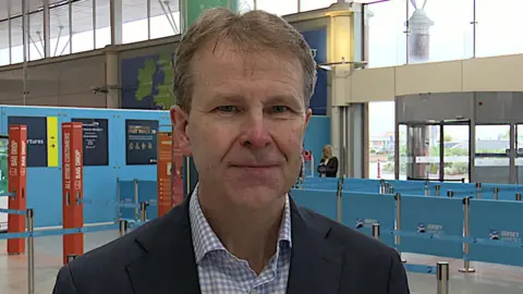 A man wearing a suit and shirt standing in front of the indoor entrance to departures at Jersey Airport.