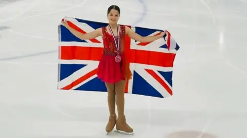 BRITISH ICE SKATING Leilah is on the ice with a British flag held behind her back. She is smiling. 