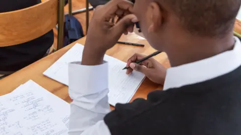 Getty Images A boy sitting in school at a wooden table. He is writing on a sheet of paper with a pencil. He is wearing a white shirt and black jumper.