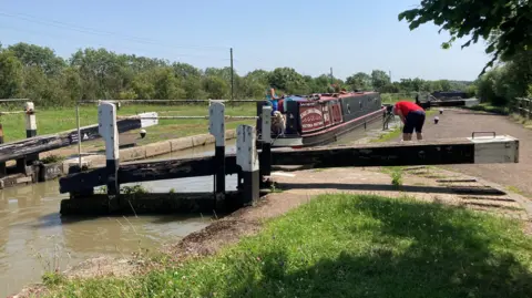 A canal lock, with a man bending over, water to the left and grass to the right. Trees are in the distance. A narrowboat is on the water, with a dog and a man at the front.
