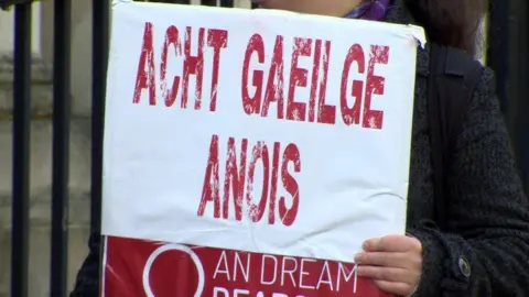 A sign reading 'Acht Gaeilge Anois' in red text on a white background, a hand can be seen holding it in front of a person wearing a dark woollen coat