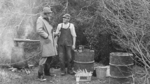Fyfe Robertson stands with a man in front of barrels used for poteen.