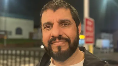 A man with a beard smiles in the dark. Road signs are illuminated behind him.