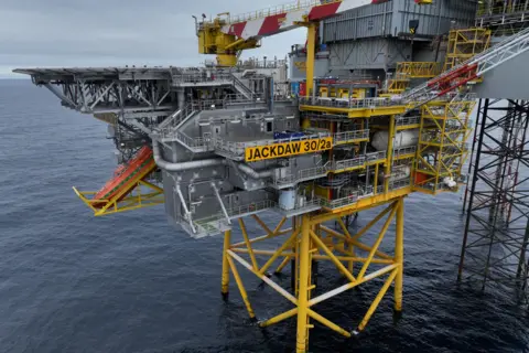 Offshore oil and gas platform marked “JACKDAW 30/2a” standing on yellow support legs above the sea, with industrial equipment, pipework and a crane visible under a partly cloudy sky.