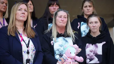 PA Media A group of women, some wearing black hooded sweatshirts with a picture of a nine-year-old girl printed onto the front. One of the women is holding a pink teddy-bear.