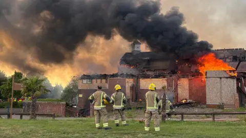 A golf club building with boarded up windows with smoke billowing upwards and part of the roof destroyed.