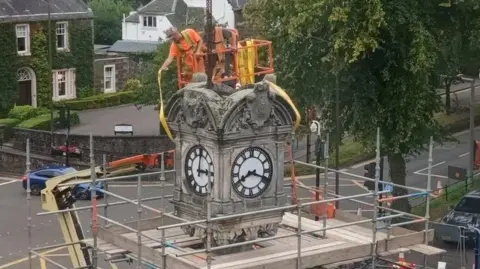 A clock tower surrounded by scaffolding with two men in orange hi vis and hard hats working on the top from a cherrypicker.