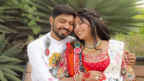 A picture of Vaibhav Patel and Jinal Goswami hugging. He is wearing a white outfit and she is wearing a red and white traditional dress.