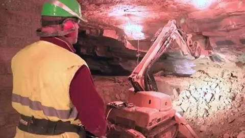 A man in yellow hi-vis and a green hard hat is operating a machine which is cutting stone. It has a long arm which is working the stone.