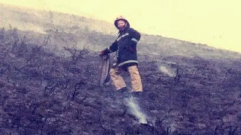 Steve Paige An older photo of Steve in a black firefighting jacket ad brown firefighting trousers. He wears a yellow helmet and is pointing towards scorched earth on a hill that has smoke coming from it