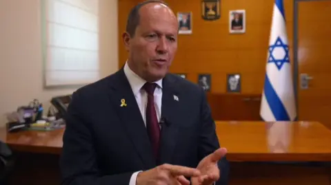Nir Barkat speaking in an office, behind him is a large wooden desk and Israel's flag.