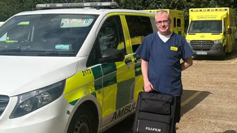 A man stood next to a van in a yellow and green ambulance livery. He is dark blue hospital scrubs and is holding a black backpack on top of a black plastic carry case.