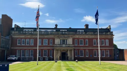 A thee-storey large building with red brick and a stone entrance. There is grass in front of it with two flag poles, one the union flag and one with Northamptonshire Police logo.