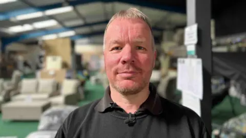 BBC/Harry Parkhill A man with short grey hair and a stubbly beard looks at the camera. He's wearing a black jumper. In the background are blurred out garden furniture items on display in a garden centre.