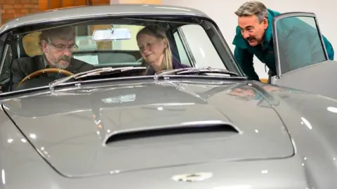Dominic Fraser/Aston Martin Works John and Susan sit in the silver birch grey car looking at the dashboard as an Aston Martin employee smiles at them from outside the car