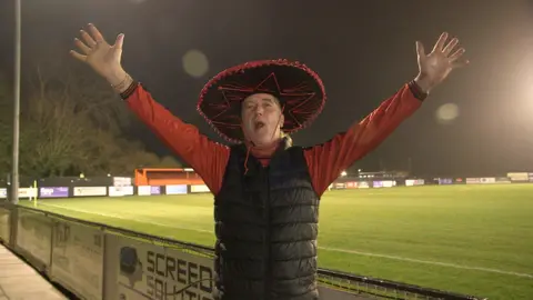 Clive Miller stands in front of the pitch wearing a red top, black gilet and a sombrero.