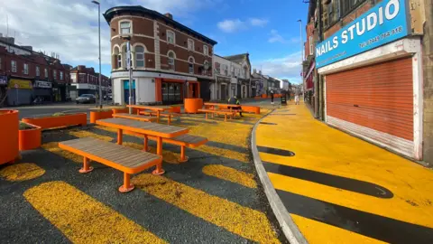 LDRS Yellow elongated markings of different sizes are seen on the tarmac on a street with the reverse pattern of black elongated markings of different lengths on a yellow pavement. Orange benches and planters are also placed along a section of the road.