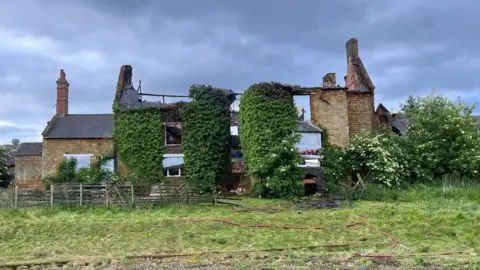 A burnt out shell of a farmhouse building. The roof of the structure has completely collapsed as a result of fire.