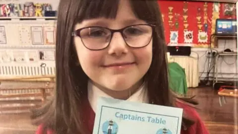 A young girl at school in a red jumper and white shirt, she has dark hair and a fringe and glasses and holds an award which says 'capain's table'
