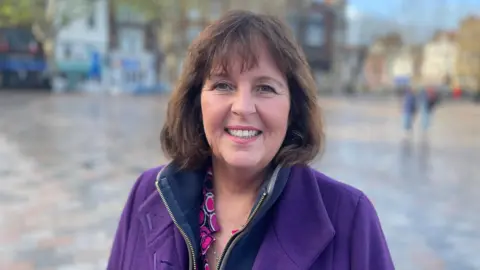 Lucy Beck from Salisbury's Chamber of Commerce stands in the centre of the city's Market Square. She's wearing a purple coat, she has dark hair with a fringe, and she is smiling