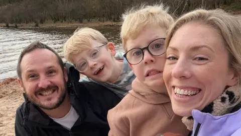 Daisy Woods Daisy Woods, pictured right, smiles in a selfie with her husband Andy (left), son Oscar (second left) and son Sonny (second right). They are on a beach with trees in the background and all are wearing coats and fleeces.