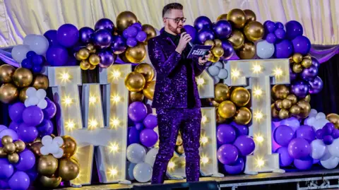 James Clarke, dressed in a purple glitter suit, on stage during Worksop's Got Talent, against a backdrop of colourful balloons and the illuminated letters WGT