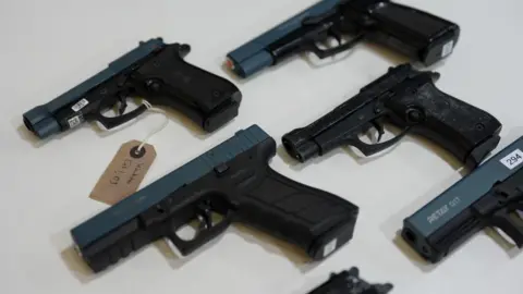 Five black and blue handguns lie flat on a cream table. One has an evidence tag tied to it.