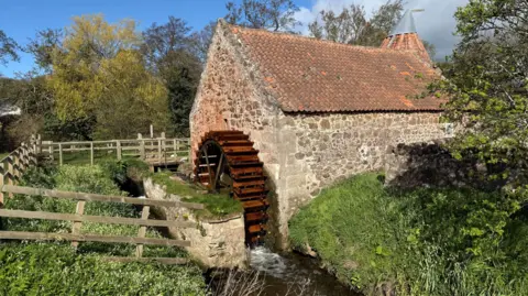 PA Media A waterwheel sits in a burn and is attached to a red brick building with a red tiled roof.