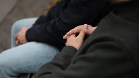 A close-up of two men sitting on what appears to be a park bench. Their faces are not visible but we see the hands of one of the men clasped together.