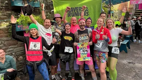 Ravers2Runners A group of 10 runners in various bright coloured items of clothing, ranging form neon tights and bra under a top to pink shorts and bright coloured hats. They are all smiling at the camer and standing in front of a Ravers 2 Runners neon green banner and holding a leaflet for Dry Run. There are people milling around behind them. 